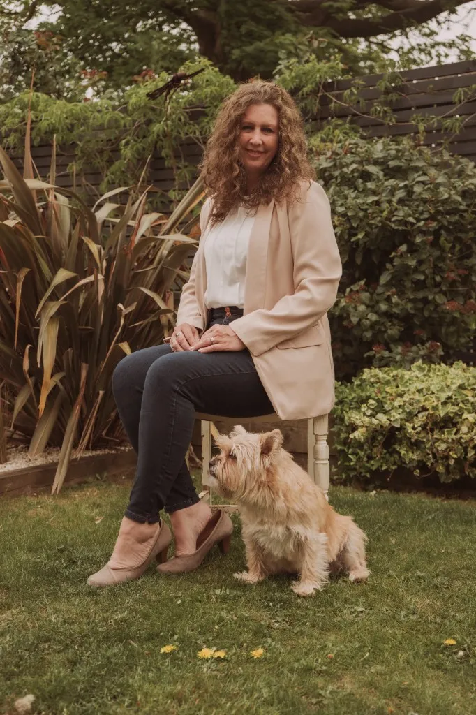 Ally seated on a wooden stool in her garden with her cairn terrier sitting attentively beside her