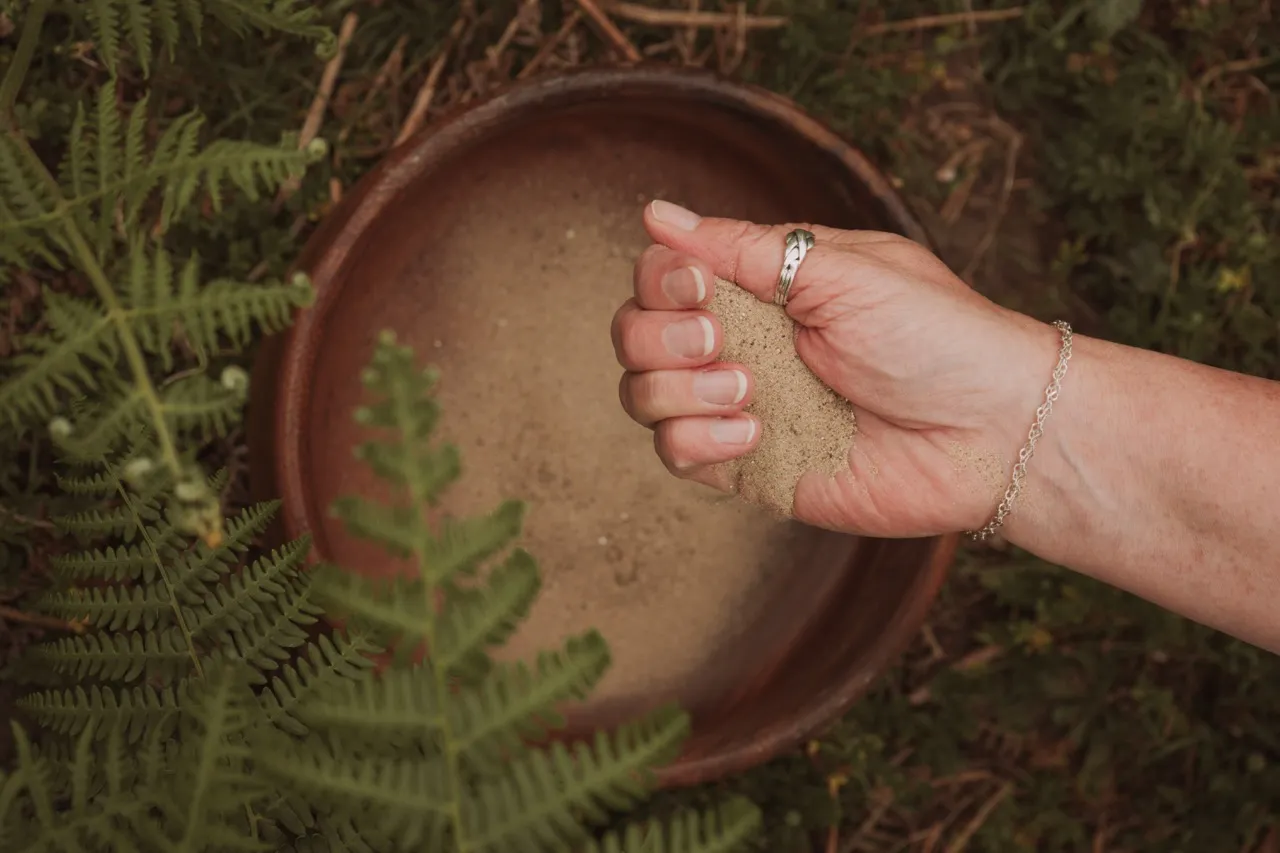 A hand gently letting sand fall into a terracotta bowl surrounded by ferns — a grounded, reflective image evoking presence and quiet connection.