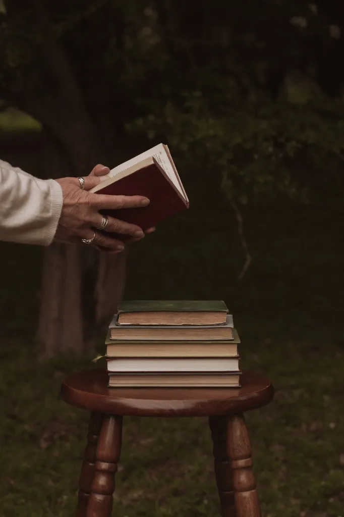 A hand gently lifting a hard-backed book from a stack on a wooden stool in a quiet woodland — choosing what to carry, what to put down