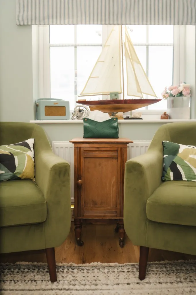 Two soft green velvet armchairs facing each other in Ally's therapy room, with a small wooden cabinet between them — a calm space for couples sessions