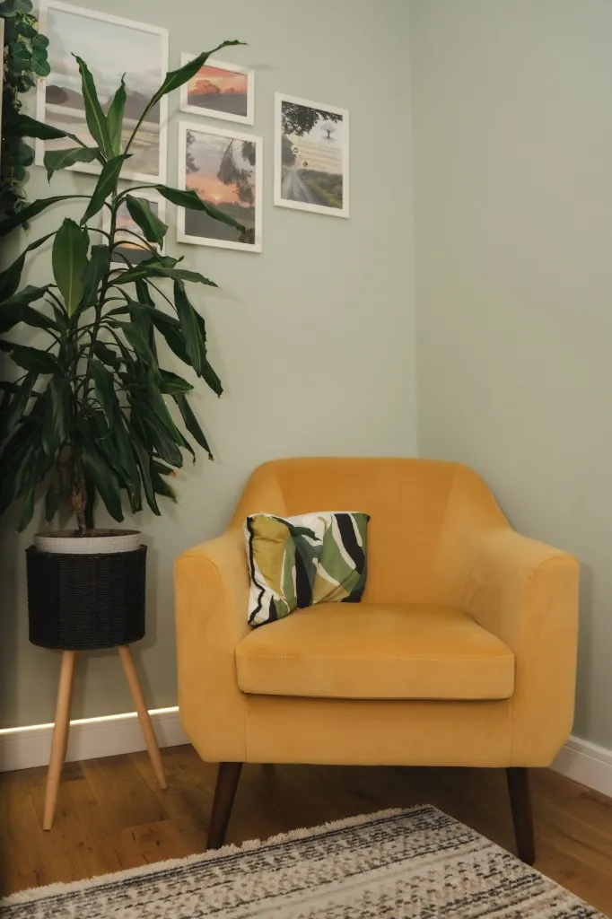 A soft mustard armchair against a sage-green wall in Ally's therapy room, with framed landscape photographs above and a leafy plant alongside — the individual session space