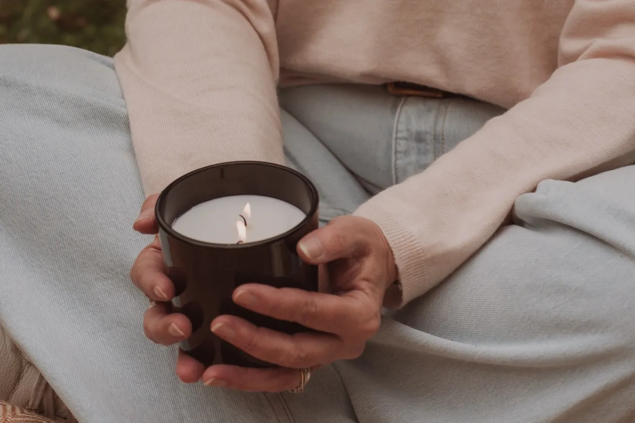 Hands holding a lit candle while sitting quietly in soft pastel light — a grounding pause that invites noticing emotional patterns.