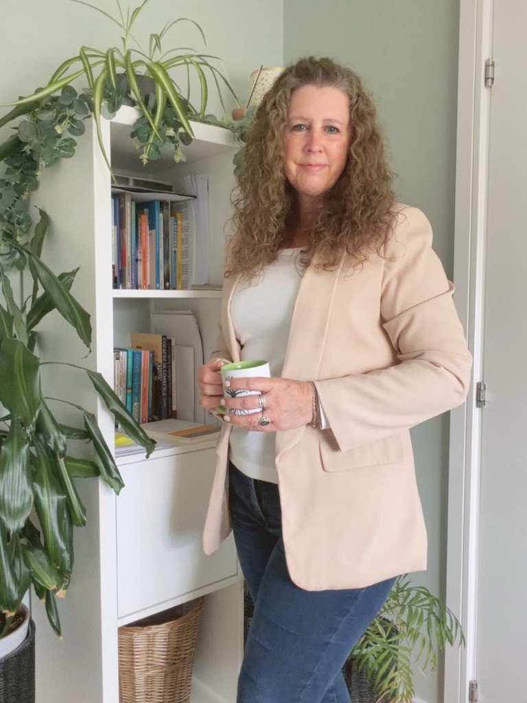 Ally standing by a plant-filled bookshelf in her therapy room, holding a sage mug